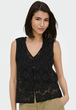Femme aux cheveux foncés mi-longs portant un haut en dentelle noir sans manches avec des boutons et un pantalon beige, debout avec une main dans la poche.