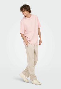 Young man smiling, wearing a loose pink t-shirt, beige pants, white sneakers, hands in pockets, against a plain light background.