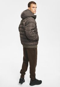 Brązowa pikowana kurtka puchowa z kapturem, z poziomymi przeszyciami i ściągaczami na rękawach, w połączeniu z ciemnobrązowymi spodniami dresowymi i czarnymi butami.