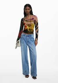 Langærmet top med et farverigt blomster- og geometrisk mønster, parret med brede blå denimjeans. Accessories inkluderer en lys håndtaske.