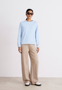 Világoskék kapucnis felső, domború logóval, bézs, bő szárú nadrággal párosítva. A modell oversize sötét napszemüveget visel, és egy semleges színű padlón áll.