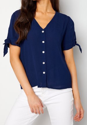 Femme portant un chemisier bleu marine à manches courtes avec des boutons sur le devant et des manches nouées, associé à un pantalon blanc.