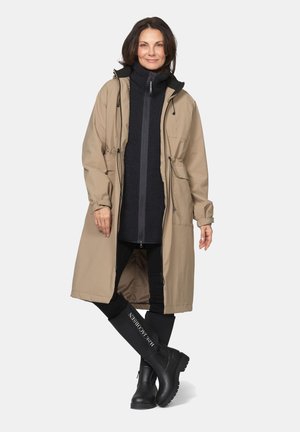 Woman wearing a beige long coat, black zip-up jacket, black leggings, and black knee-high boots standing with legs crossed.