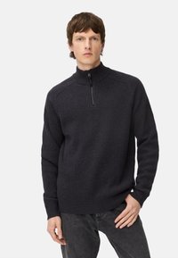 Mørkegrå lynlåstrøje i ribstrikket stof med høj krave og lange ærmer, kombineret med mørke jeans. Ren, moderne design.