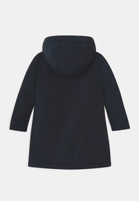 Námorně modrý kabát s kapucí a dlouhými rukávy, hladká textilie a elastické manžety. Má lem, který sahá k polovině stehna.