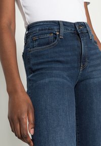 Jean en denim bleu foncé à taille haute, avec un design classique à cinq poches et des accents de couture subtils le long des coutures.