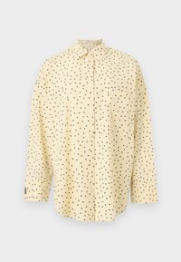 Chemise beige à manches longues avec des pois noirs et une poche poitrine sur un fond uni.