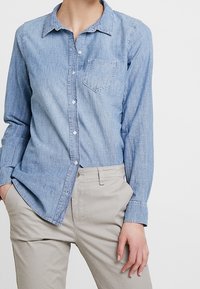 Femme portant une chemise en denim bleu clair avec des boutons et un pantalon beige, les mains dans les poches, se tenant devant un fond blanc uni.
