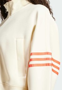 Krämfärgad tröja med hög krage, med en orange-randig detalj på ärmen och en liten ficka framtill.