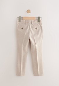 Pantalon beige sur mesure suspendu à un cintre en bois avec des pinces métalliques, vu de dos avec deux poches passepoilées boutonnées.