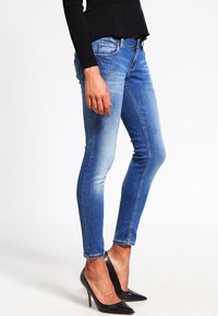 Jeans skinny de mezclilla azul con un acabado desgastado, con un talle medio y un diseño clásico de cinco bolsillos, combinados con tacones stilettos negros.