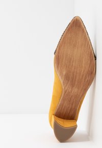 Scarpa con tacco alto in suede giallo vista dal basso, con una suola in legno caratterizzata da un design testurizzato e un tacco curvato.