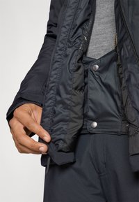 Crna izolirana jakna s glatkom vanjštinom, unutarnja podstava vidljiva, s kopčanjem na dugmadima i sivoj tkanini ispod. Nema vidljivih uzoraka.