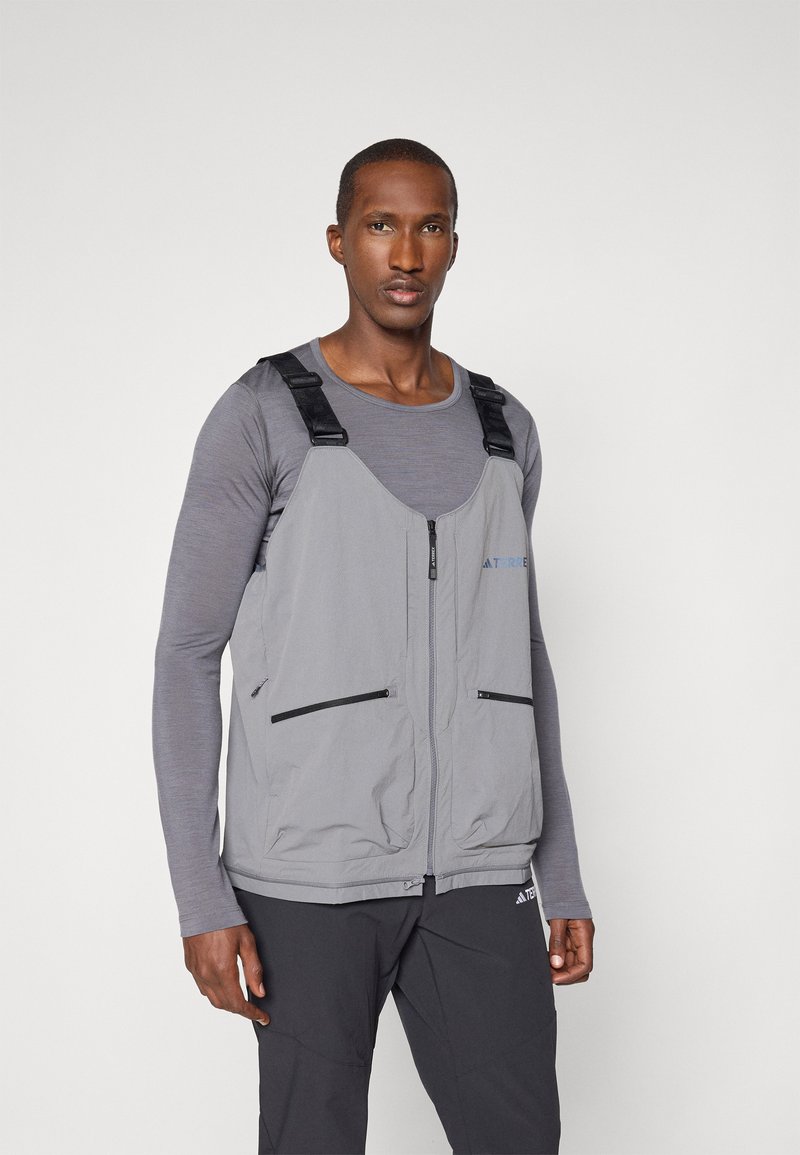 Gray vest with adjustable black straps, zippered pockets, and a smooth texture. Worn over a gray long-sleeve shirt, showcasing a minimalist design.