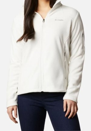 Frau trägt eine weiße Columbia-Fleecejacke mit durchgehendem Frontreißverschluss, Reißverschlusstaschen an den Seiten und einer Reißverschlusstasche am Ärmel, kombiniert mit dunklen Jeans.