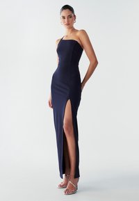 Tamsiai mėlyna, prigludusi, be petnešėlių maksi suknelė su aukštu šoniniu iškirpimu. Lygi tekstūra ir švarus, minimalistinis dizainas. Papuošta sidabrinėmis aukštakulnėmis.