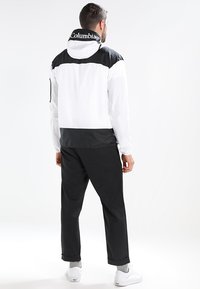 Mand i en sort og hvid Columbia-jakke, sorte bukser, grå sokker og hvide sneakers, der står med ryggen til på en hvid baggrund.