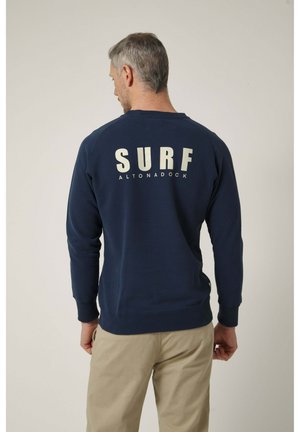 Hombre con cabello gris que lleva un sudadera azul marino con el texto "SURF ALTONADOCK" en la espalda y pantalones beige, mirando de espaldas a la cámara.