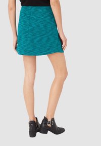 Mujer vistiendo una falda texturizada azul y botas negras tobilleras con hebillas, vista desde atrás sobre un fondo claro y liso.