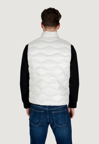 Hvid, kviltet puffer vest med høj krave, der har en shiny overflade. Bærer den over en sort langærmet skjorte og kombinerer den med blå jeans.