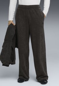 Mørkegrå sweatpants med elastisk talje, sidelommer og en diskret tekstureret overflade. En matchende jakke holdes i den ene hånd.