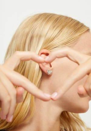 Mujer rubia formando un corazón con los dedos que enmarcan un pendiente plateado en forma de corazón en su oreja izquierda sobre un fondo blanco.