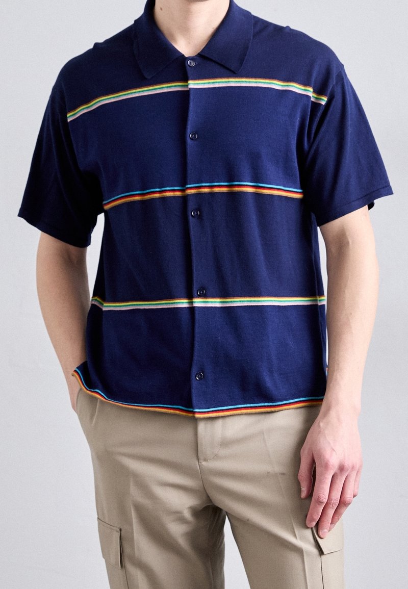 Homme portant une chemise à manches courtes bleu marine à boutons avec des rayures horizontales multicolores et un pantalon cargo beige.