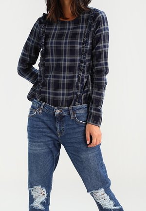 Nainen, jolla on yllään laivastonsininen ja harmaa ruudullinen pitkähihainen yläosa, jossa röyhelödetaljeja, yhdistettynä sinisiin revittyihin farkkuihin ja rentoon asentoon.