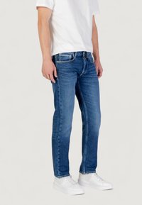 Jeans in denim blu con taglio dritto, dotati di cinque tasche, apertura con zip e leggero sbiadimento lungo le gambe. Indossati con una maglietta bianca e sneaker.