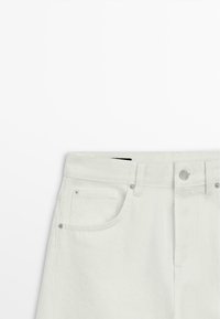 Zone de la taille d’un jean en denim blanc montrant un bouton avant, des passants de ceinture et une poche avant avec des détails de rivets sur un fond blanc uni.