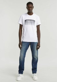 Camiseta blanca de manga corta con un estampado gráfico en tonos grises en el centro, combinada con jeans azules y zapatillas blancas.