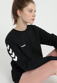 Jonge vrouw met paardenstaart zittend, draagt een zwart shirt met lange mouwen met witte chevronstrepen en lichtroze getextureerde legging.