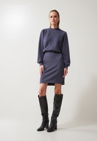 Robe longue à manches longues gris foncé avec une silhouette ajustée, présentant des épaules bouffantes. Une paire de bottes à talons hautes en cuir noir complète le look.