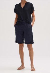 Vrouw in een zwarte korte mouwen shirt met structuur, marineblauwe opgerolde shorts met koordje, handen in de zakken, en beige platte sandalen.