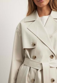 Smėlinis dvigubas trenčkotas, pagamintas iš audinio, su dideliais mygtukais, įpjovomis apykaklėmis ir derančiu diržu reguliavimui.