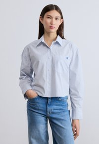 Ριγέ πουκάμισο σε ανοιχτό μπλε χρώμα με κουμπιά, τσέπη στο στήθος, άνετη εφαρμογή και μακριά μανίκια, συνδυασμένο με μπλε τζιν. Διακριτικό λογότυπο στην τσέπη του στήθους.