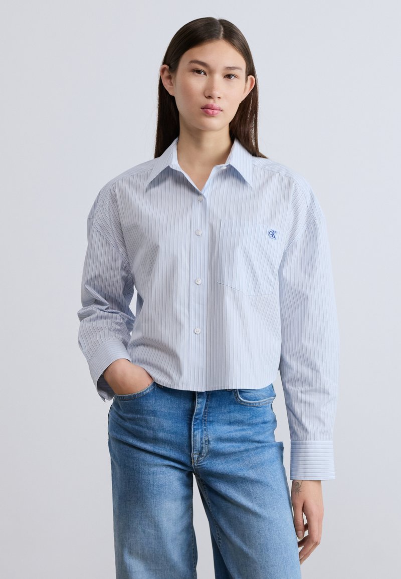 Ριγέ πουκάμισο σε ανοιχτό μπλε χρώμα με κουμπιά, τσέπη στο στήθος, άνετη εφαρμογή και μακριά μανίκια, συνδυασμένο με μπλε τζιν. Διακριτικό λογότυπο στην τσέπη του στήθους.