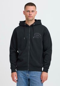 Sort lynlås hoodie med foret hætte, snore, frontlogo og ribbede manchetter. Bæres over blå jeans og har en glat overflade.