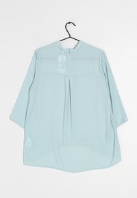 Blouse bleu clair à manches longues en tissu doux, avec un col rond, des détails de plis à l'avant et une petite fermeture éclair à l'arrière.
