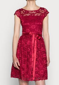 Robe rouge en dentelle avec manches courtes, corsage ajusté et jupe évasée. Dotée d'une ceinture en satin et d'un détail nœud. Ourlet festonné.