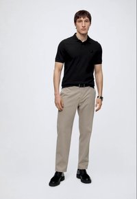 Hombre con una camiseta polo negra, pantalones beige con cinturón, zapatos negros gruesos y un reloj de pulsera, de pie frente a un fondo liso y claro.