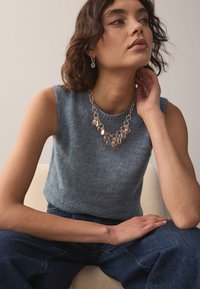 Gray knitted sleeveless top with a round neckline, paired with a multi-layered gold necklace featuring blue and pink accents. Dark denim jeans.