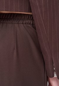 Close-up van een persoon die donkerbruine elastische taillebroeken draagt en een bijpassende pinstripe blazer met een gespleten manchet.