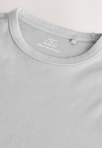 Harmaa ribbineulospaita, jossa pyöreä pääntie. Kevyt kangas, jossa hienovaraiset pystysuorat viivat. Sisäpuolella oleva merkintä: "Next made in Bangladesh."