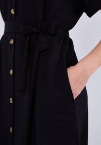 Abito nero con bottoni e maniche corte, dotato di una cintura annodata in vita, tasche laterali e una superficie liscia con un leggero lucido.