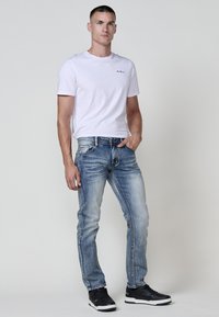 Hombre de pie con la mano en el bolsillo, vistiendo una camiseta blanca ajustada, pantalones vaqueros ajustados de color azul desgastado y zapatillas negras, contra un fondo liso.