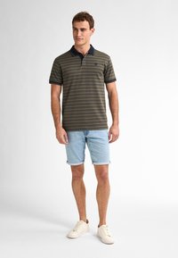 Polo tričko s krátkým rukávem v olivově zelené barvě s námořnickými pruhy, kombinované se světle modrými denimovými šortkami. Model ve bílých teniskách stojí na neutrálním pozadí.