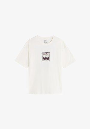 White cotton t-shirt featuring a central black graphic of scales within a decorative border. Short sleeves and a crew neckline.