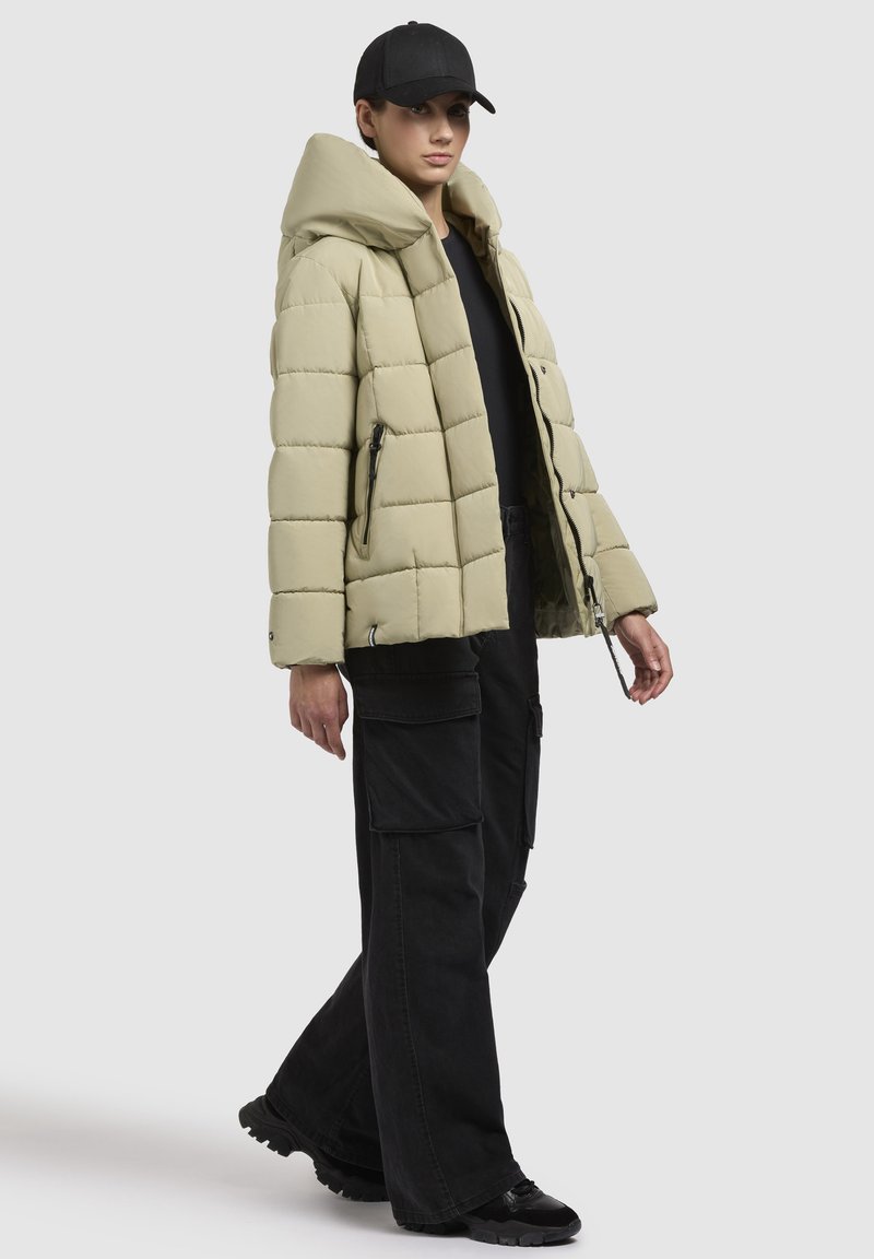 Giacca puffer beige chiaro con cappuccio, caratterizzata da un motivo trapuntato e tasche laterali con zip. Abbinata a pantaloni cargo neri e scarpe sportive.
