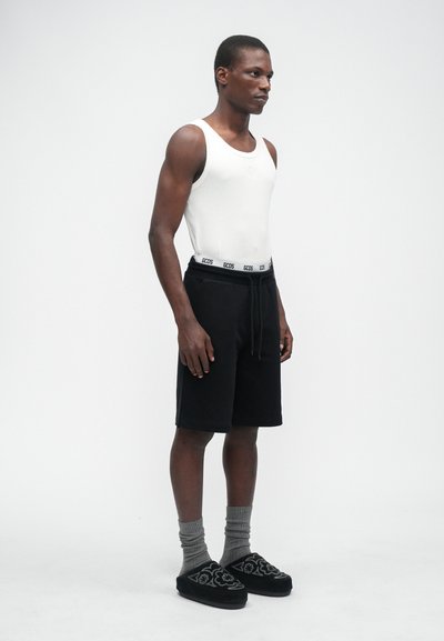 Weißes Tanktop, schwarze Shorts mit Kordelzug, graue Socken, schwarze Schuhe mit Muster, stehend gegen einen einfarbigen Hintergrund.